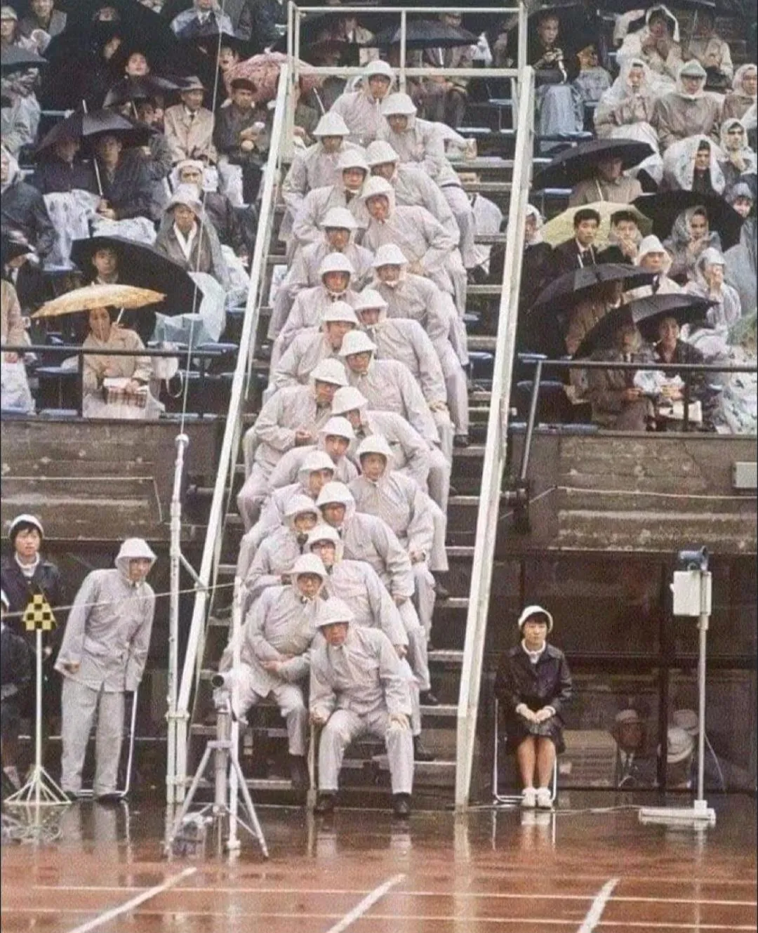 Rows of uniformed officials seated along the finish line at the 1964 Tokyo Olympics, umbrellas up in the rain.
